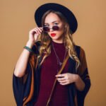 studio-close-up-portrait-young-fresh-blonde-woman-brown-straw-poncho-wool-black-trendy-hat-round-glasses-looking-camera-green-leather-had-bag